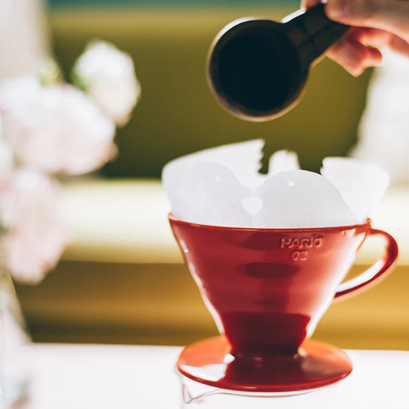 Nærbillede af en rød kaffedrypper med et filterpapir indeni. En hånd holder en sort måleske over drypperen. Baggrunden er blødt sløret og viser en grøn sofa og nogle lyserøde blomster.