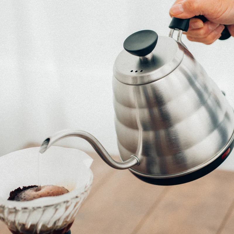 En hånd hælder varmt vand fra en svanehalskedel i rustfrit stål i et kaffefilter med malet kaffe ved hjælp af en hældningsmetode til brygning.