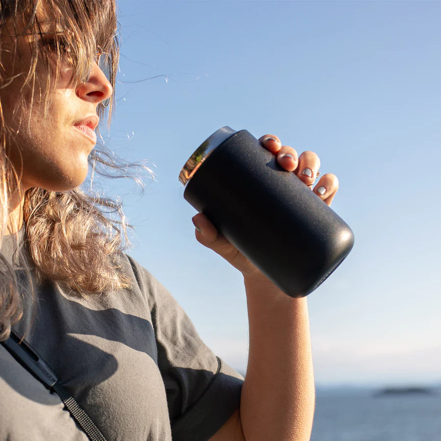 En person med skulderlangt hår og solbriller drikker af en sort isoleret glas på en solskinsdag med en klar blå himmel og et glimt af havet i baggrunden.