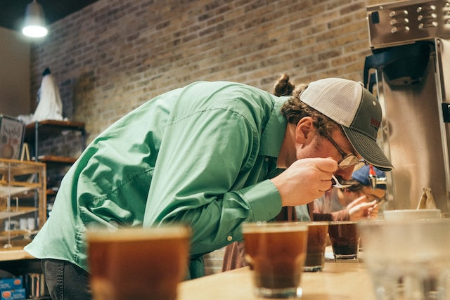 En person i en grøn skjorte og kasket læner sig ind over et bord for at smage kaffe fra en ske, og deltager i en kaffekoppesession inde i en hyggelig café med murstensvægge i baggrunden.