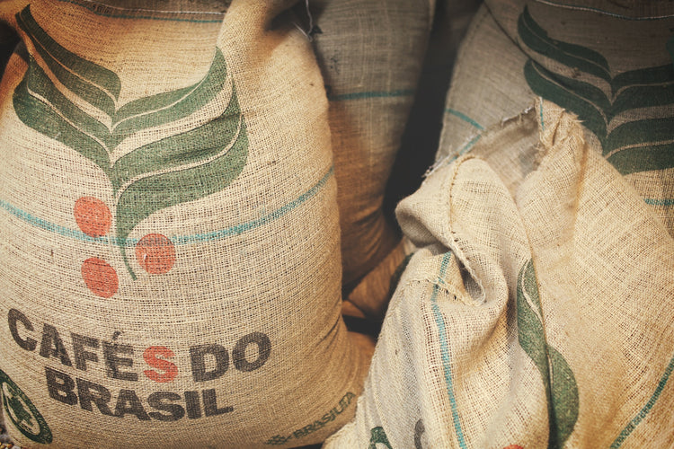 Jutesække fyldt med kaffebønner mærket "Cafés do Brasil." Sækkene viser grønne og røde kaffeplanter, der angiver oprindelsen af kaffebønnerne fra Brasilien.