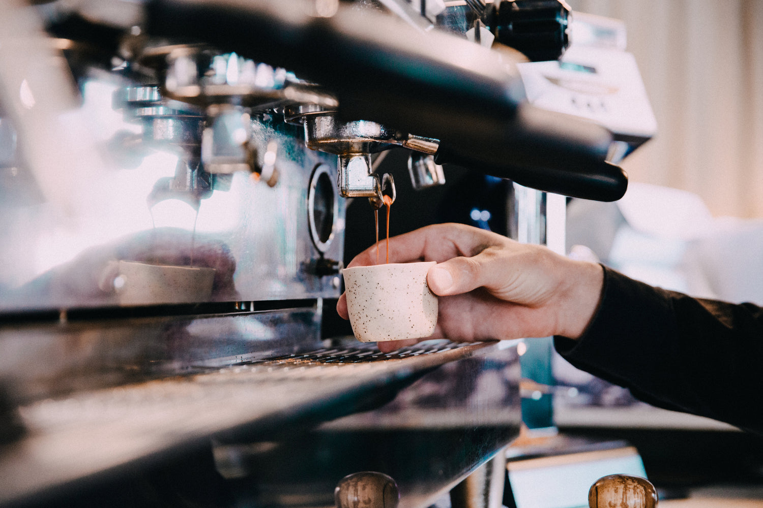 En person holder en lille kop under tuden på en skinnende espressomaskine, mens kaffe drypper ned i koppen. Personens hånd er synlig og styrer koppen for at fange kaffen. Baggrunden er lidt sløret.