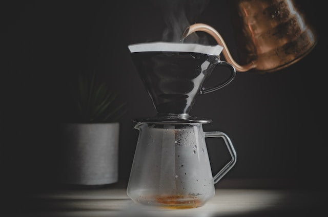 Et nærbillede af en kaffebrygningssetup med en hæld-over-drypper placeret oven på en glaskaraffel. Vand hældes fra en kobberkedel i drypperen. Der stiger damp fra kaffebryggen. En lille potteplante er sløret i baggrunden.