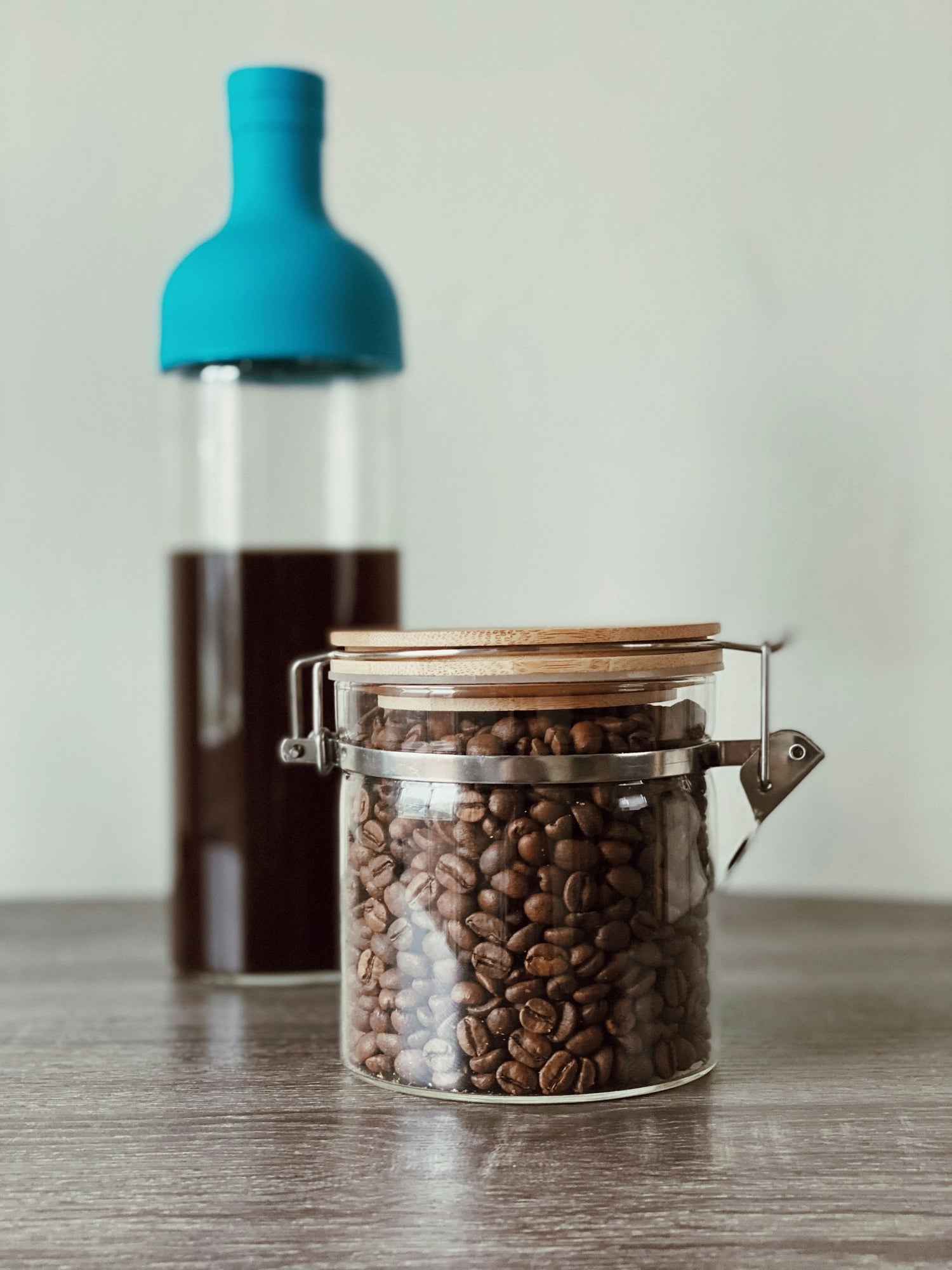 En klar glaskrukke fyldt med kaffebønner, forseglet med et trælåg, står på en træoverflade. I baggrunden er der en høj flaske med en blå top, der indeholder mørk væske, sandsynligvis koldbrygget kaffe. Baggrunden er en almindelig lysegrå.