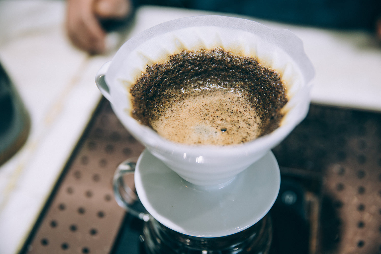 Nærbillede af en pour-over kaffe i en hvid keramisk drypper med papirfilter. Kaffegrumset blomstrer, synligt med bobler og skumdannelse. Drypperen hviler oven på en glaskaraffel. En persons hånd ses i den slørede baggrund.