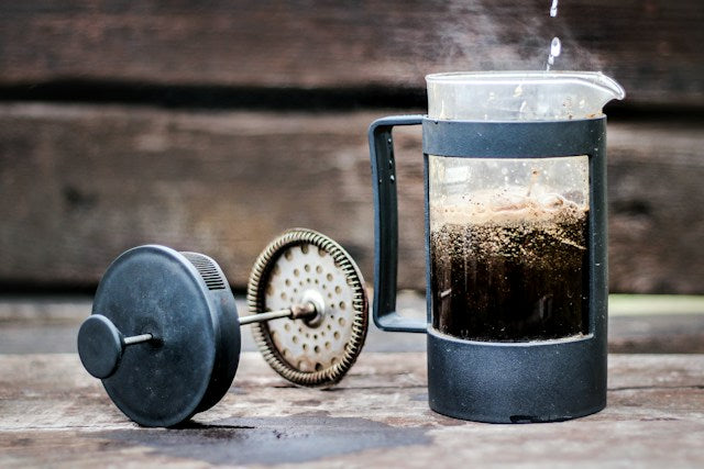 En sort fransk presse med kaffegrums indeni sidder på en træoverflade. Låget og stemplet sættes ved siden af, og dampen stiger op fra kaffen. Baggrunden har mørke træpaneler.