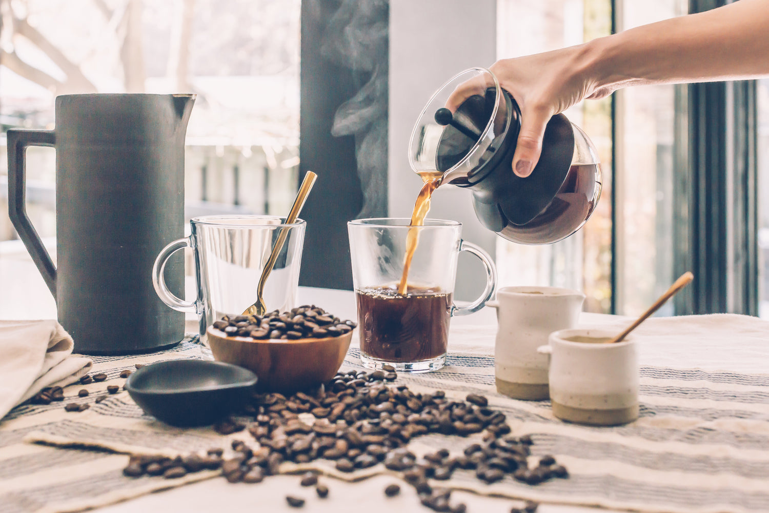 En hånd hælder kaffe fra en glaskande i et glaskrus på et bord. Spredte kaffebønner, en sort skål, to små kopper, en kande og en ske står også på bordet. Damp stiger fra kruset, hvilket indikerer varm kaffe.