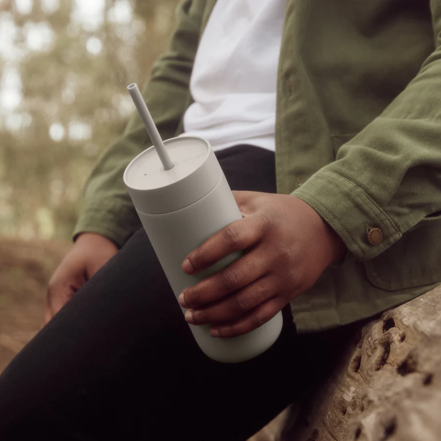 En person i en grøn jakke holder en Fellow Carter Cold Tumbler, ICED Coffee rejse krus, mens han sidder på en træstamme udendørs. Den slørede baggrund antyder rolige naturlige omgivelser, ideel til at nyde øjeblikket.