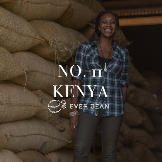 En smilende person står ved siden af store stakke af jute-sække fyldt med EverBeans nr. 11 · Kenya-kaffe, kendt for sine rige espresso-noter. Tekstoverlejringen lyder "NR. 11 KENYA," ledsaget af et subtilt logo på baggrund af en travl lagerbygning.