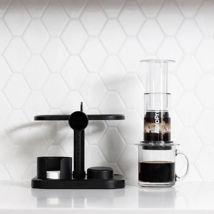 En slank AeroPress Organizer-opsætning på en hvid tæller med en sekskantet flisebagplade med Aeropress-mærket. Kaffemaskinen til højre brygger til et gennemsigtigt krus, mens venstre side har et moderne sort stativ med målekomponenter til effektiv kaffepresseopbevaring.