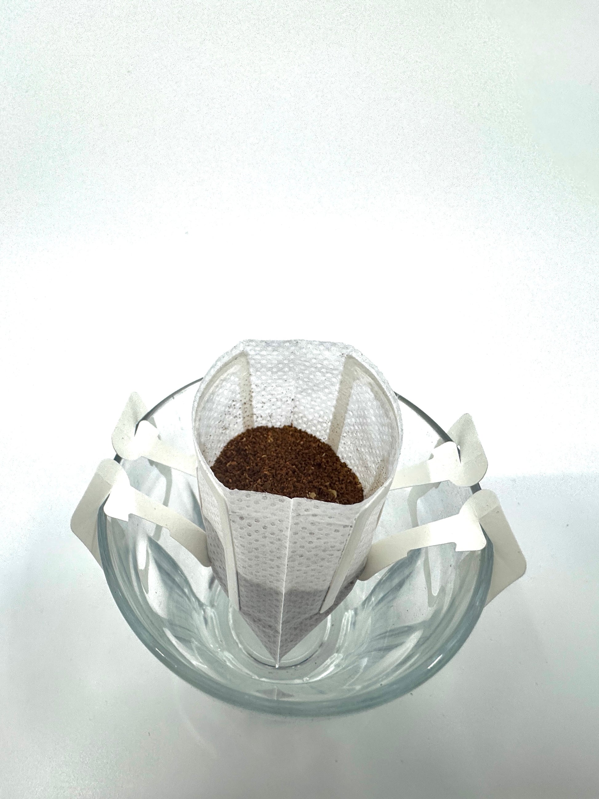 EverBeans Single Serve, colombiansk Maceration dryp kaffefilter hviler over en klar glaskop. Det hvide filter har sideflapper, der hviler på koppens kant, klar til at brygge dens unikke smag mod en almindelig hvid baggrund.