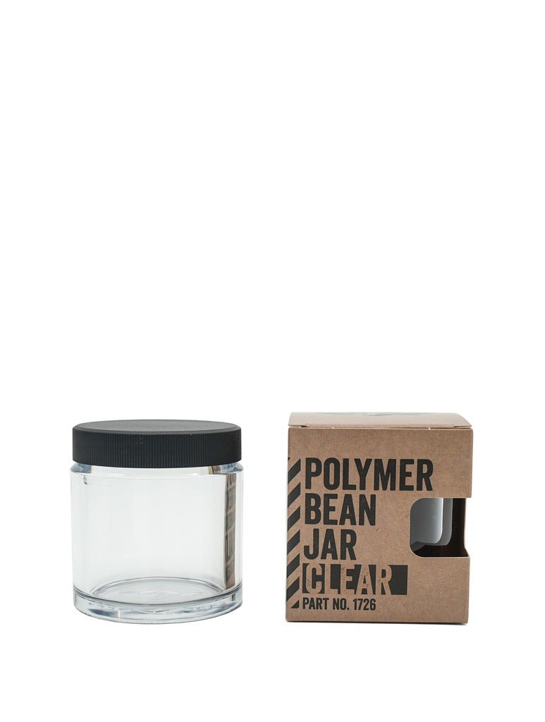 En klar cylindrisk krukke med sort låg, der minder om Comandantes Polymer Bean Jar med låg, står ved siden af en brun papkasse. Æsken er forsynet med sort tekst med teksten "Comandante Polymer Bean Jar med låg Klar", med en udskæring på den ene side, hvilket sikrer fødevaresikker-kvaliteten.