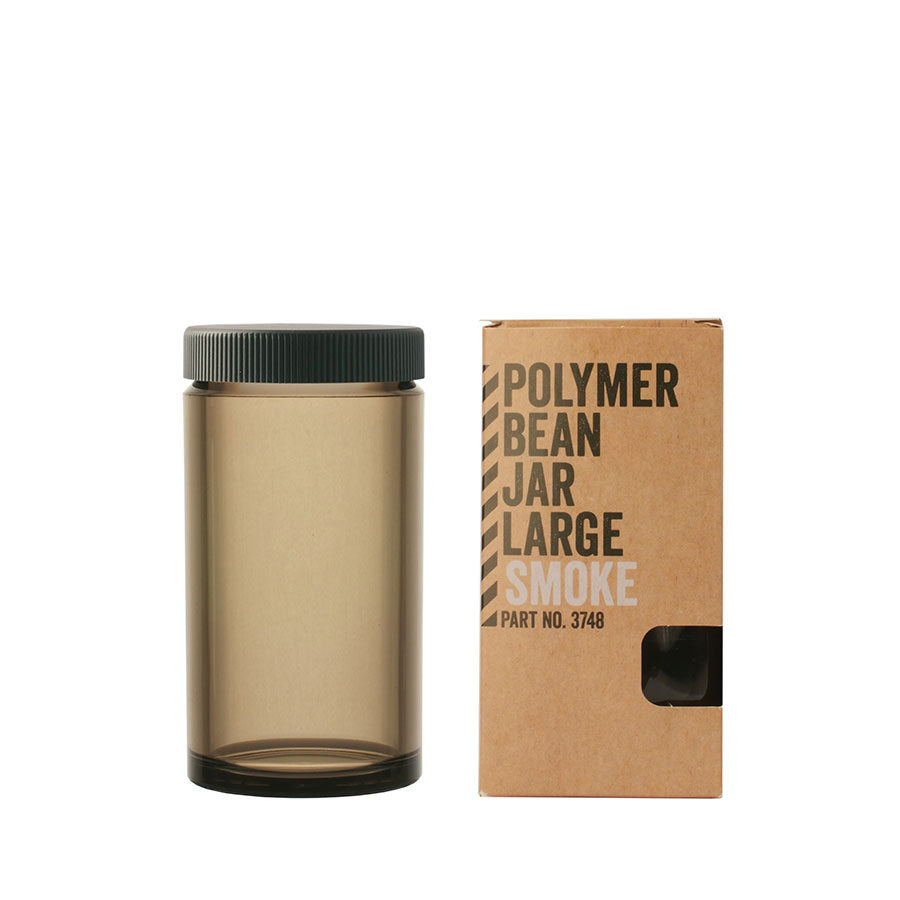 En høj, gennemskinnelig ravfarvet krukke mærket "Comandante, Polymer Bean Jar med låg, Uklar Large," med et sort låg står ved siden af en brun æske. På æsken står "Polymer Bean Jar Large Smoke" og sikrer, at det er fødevaresikker til sikker opbevaring.