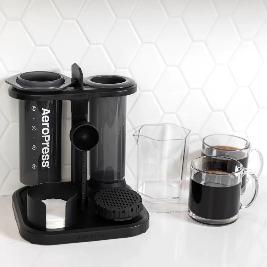 En AeroPress-enhed med to kamre sidder pænt i en AeroPress Organizer på disken sammen med en klar målekop og to klare krus sort kaffe mod en hvid sekskantet flisebelagt bagplade.