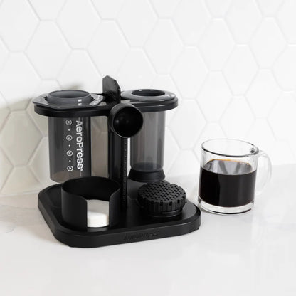 En AeroPress Organizer fra Aeropress, med to grå cylindriske kamre, en måleske og en filterholder, sidder pænt på disken. Et klart glas krus med sort kaffe sidder ved siden af det på baggrund af hvide sekskantede fliser.