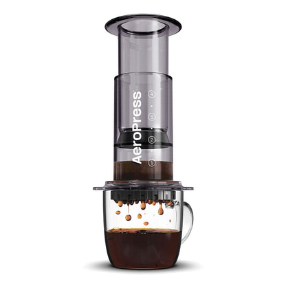 En AeroPress, Clear Kaffebrygger, Sort-limited kaffemaskine fra Aeropress hviler på et glaskrus. Dens slanke design med bryggeteknologi brygger rig kaffe, der drypper nedenunder. Den gennemsigtige enhed har karakteristiske sorte komponenter og viser stolt Aeropress-mærket.