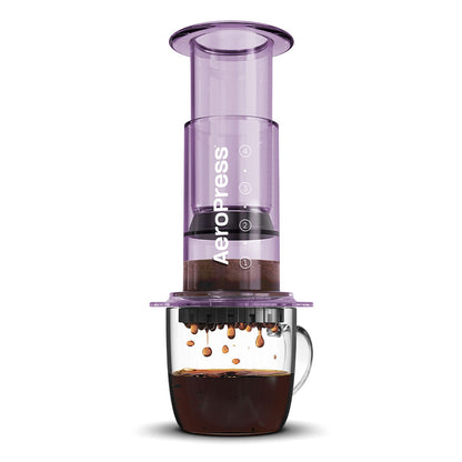 En Tritan™ gennemsigtig AeroPress, Clear Kaffebrygger Lilla-limited af AeroPress, sidder over en glaskop. Den brygger kaffe med faldende dråber og viser sin innovative bryggeteknologi og et tydeligt synligt AeroPress-logo.