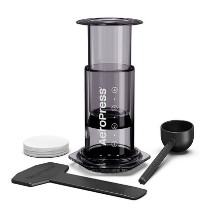 En sort AeroPress Clear Kaffebrygger, Sort-limited fra Aeropress, med en scoop, omrører og papirfiltre, er sat mod en hvid baggrund. Denne bærbare enhed fremhæver innovativ bryggeteknologi for en enestående bryggeoplevelse, uanset hvor du er.