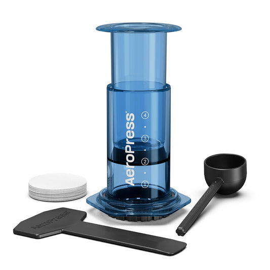 Den blå AeroPress Clear Limited kaffebrygger fra Aeropress, der er placeret på et hvidt bagtæppe, er lavet med slidstærkt Tritan™, inkluderer et stempel, et kammer, en filterhætte, en ske, omrører og papirfiltre – ideel til at forbedre din kaffeoplevelse.