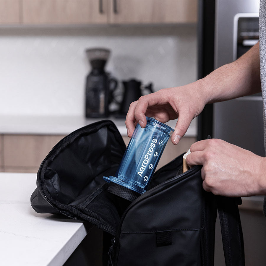 En person placerer forsigtigt en Aeropress, Clear Limited Kaffebrygger, Blå i en sort pose på en hvid køkkenbord. Træskabe og en kaffekværn forstærker kaffeoplevelsen i baggrunden.