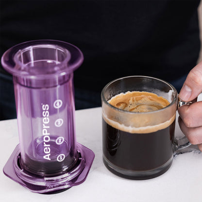 En lilla limited-edition Aeropress Clear Kaffebrygger sidder på en hvid overflade ved siden af et glaskrus fyldt med fyldig sort kaffe. En hånd griber krusets håndtag og fremhæver den overlegne "bryggeteknologi" for den perfekte bryg hver gang.