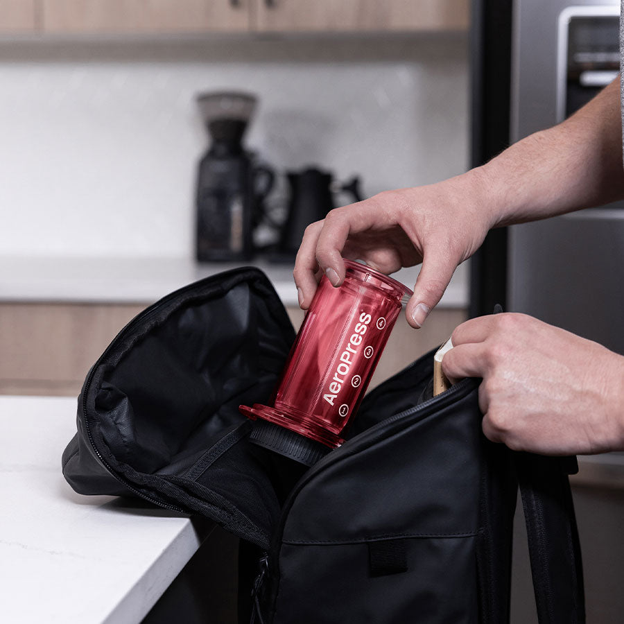 Hænderne placerer forsigtigt en rød Aeropress Clear Kaffebrygger, Rød-limited kaffemaskine i en elegant sort pose på køkkenbordet, med en kaffekværn og en kande i baggrunden.