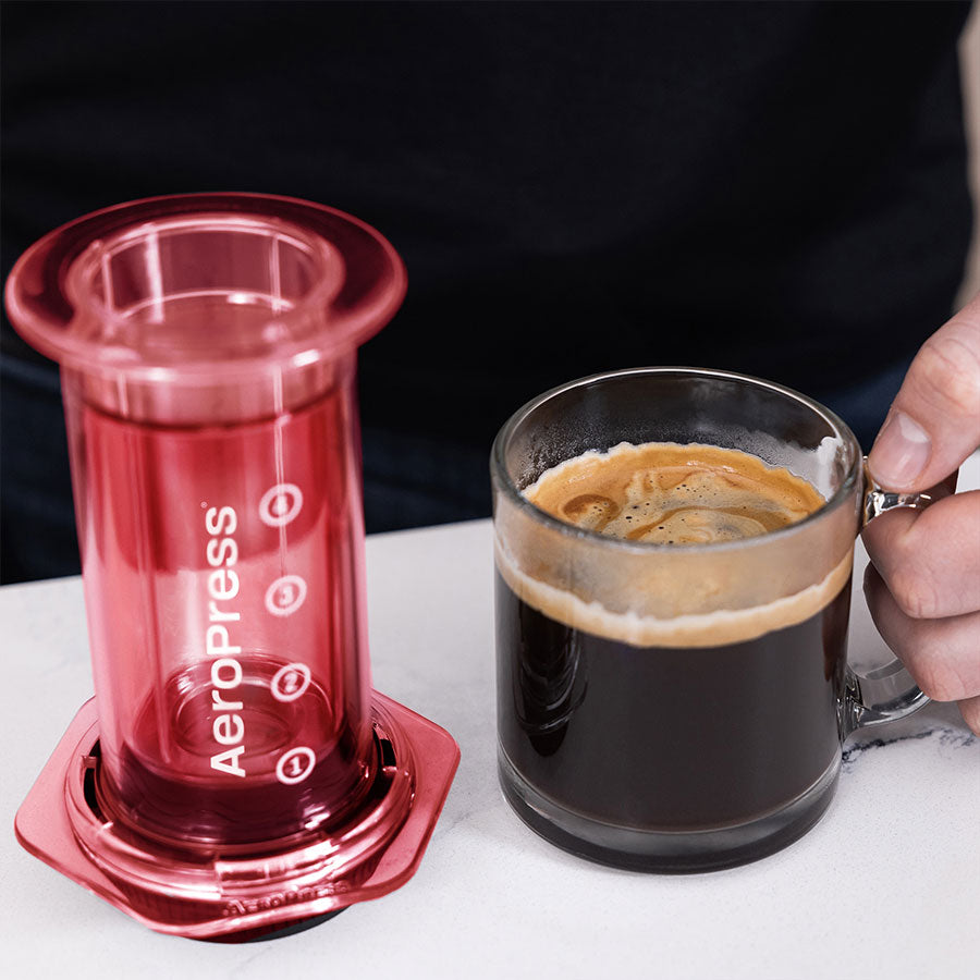 En person griber et klart Tritan™ glaskrus fyldt med sort kaffe ved siden af en rød-begrænset AeroPress, Clear Kaffebrygger fra Aeropress på en lys overflade.