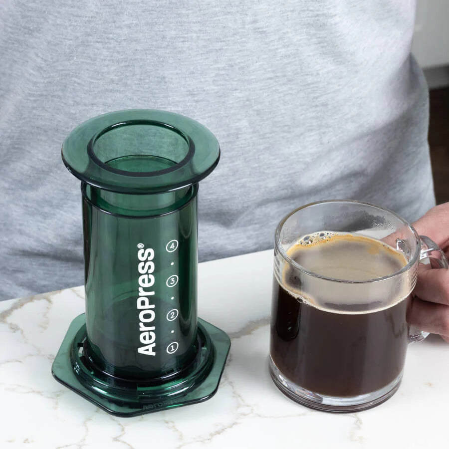 En grøn AeroPress, Clear Limited Kaffebrygger fra Aeropress, lavet af slidstærkt Tritan™, hviler på en marmorbordplade ved siden af et glaskrus med fyldig sort kaffe. I baggrunden er en person i en grå skjorte delvist synlig, hvilket forstærker den rolige morgenstemning.