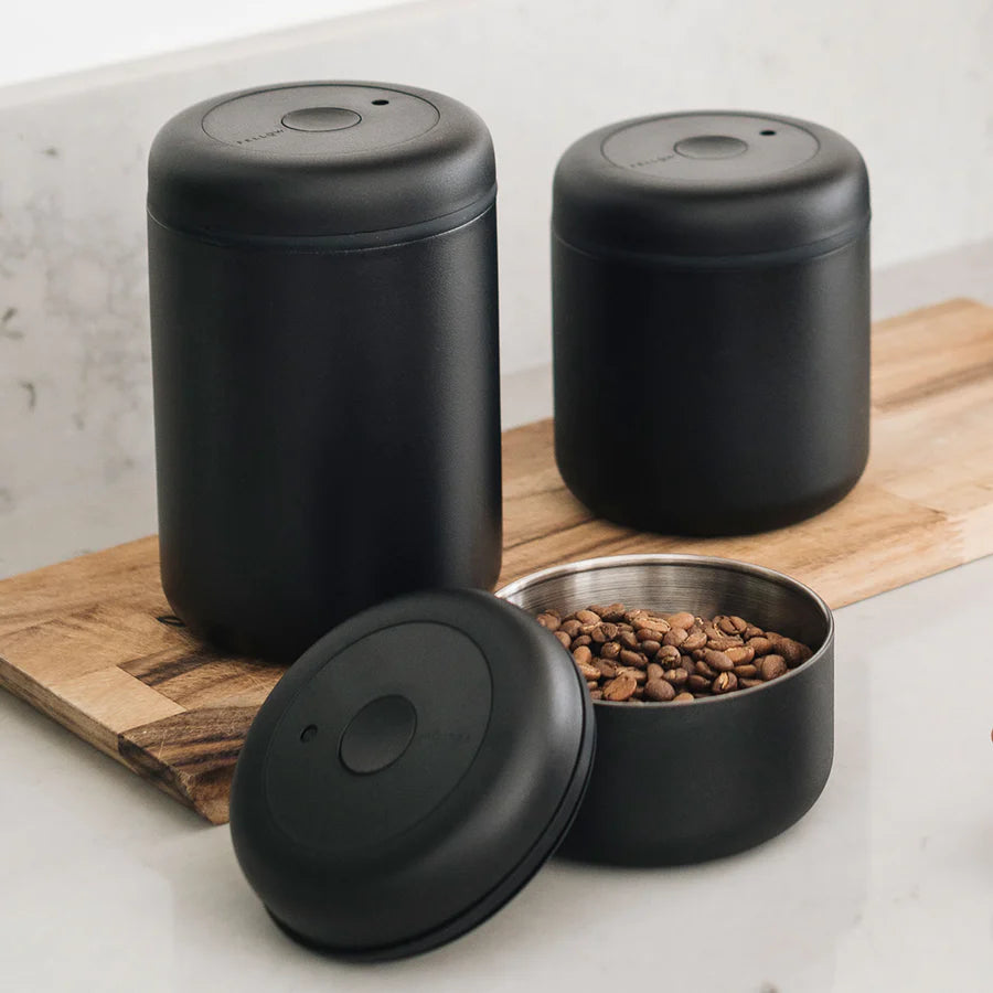 Tre sorte Fellow Atmos Vakuum Beholder-beholdere er på en træoverflade. Den ene er åben og afslører kaffebønner indeni, med vægt på opbevaring af kaffebønner. Det minimalistiske design sikrer, at de forbliver lufttæt for at forhindre oxidation, alt sammen sat mod en lys baggrund.