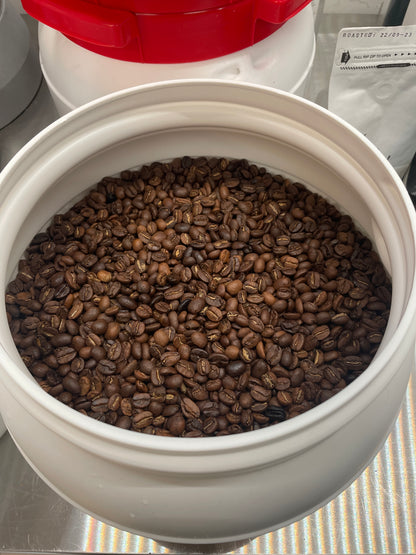 En stor hvid beholder med EverBeans nr. 06-dc, Colombia Dark Roast Decaf, fantastisk til espresso, sidder på en metaloverflade. De rige, mørkebrune bønner er smukt udstillet i den åbne beholder med en rødlåget krukke delvist synlig i baggrunden.