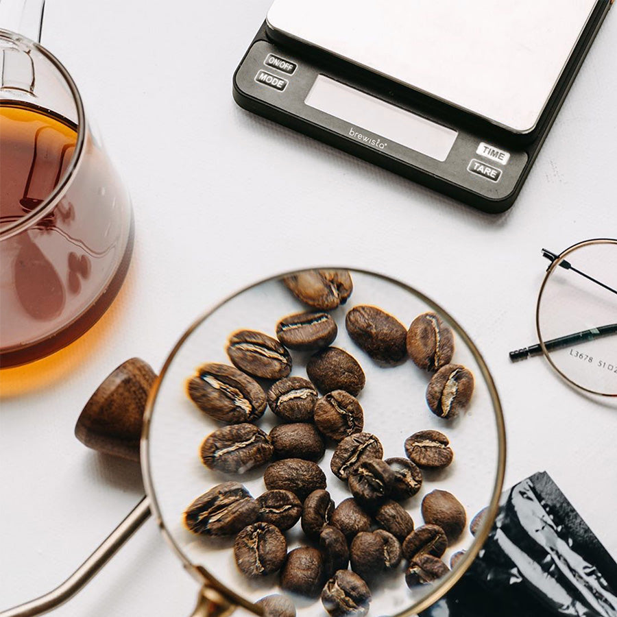 Kaffebønner forstørres på en hvid overflade. I nærheden er der en EverBean Aeropress fra Barista Pack, et glas kaffe, en Brewista kaffevægt digital vægt og et par briller.