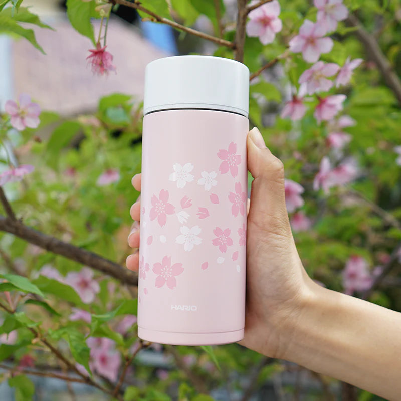 En hånd holder en pink Hario Stick-flaske 350mL Sakura, prydet med hvide og lyserøde kirsebærblomster. Baggrunden af slørede grønne blade og kirsebærblomster fremkalder en udendørs indstilling, ideel til at nyde din drink på farten.