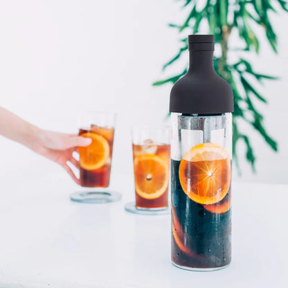 En Hario Cold Brew kaffefilter-i-flaske med mørk top rummer iste og appelsinskiver, mens to glas er fyldt med blandingen. En hånd rækker ud efter et glas ved siden af en levende grøn plante i den slørede baggrund.