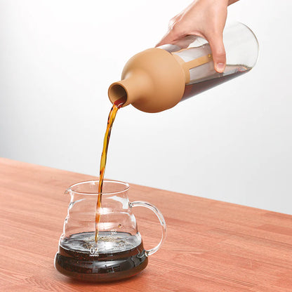 En hånd hælder koldbrygget kaffe fra Hario's Cold Brew Coffee Filter-in Bottle i en gennemsigtig karaffel på et træbord. Den mærkede Hario-karaffel indeholder lidt kaffe og hviler på en almindelig lysegrå baggrund.