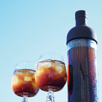 To glas iskaffe sidder ved siden af en høj, mørk Hario Cold Brew kaffefilter-i-flaske mod en klar blå himmel. Glassene er fyldt med koldt bryg og isterninger, hvilket skaber et forfriskende look.