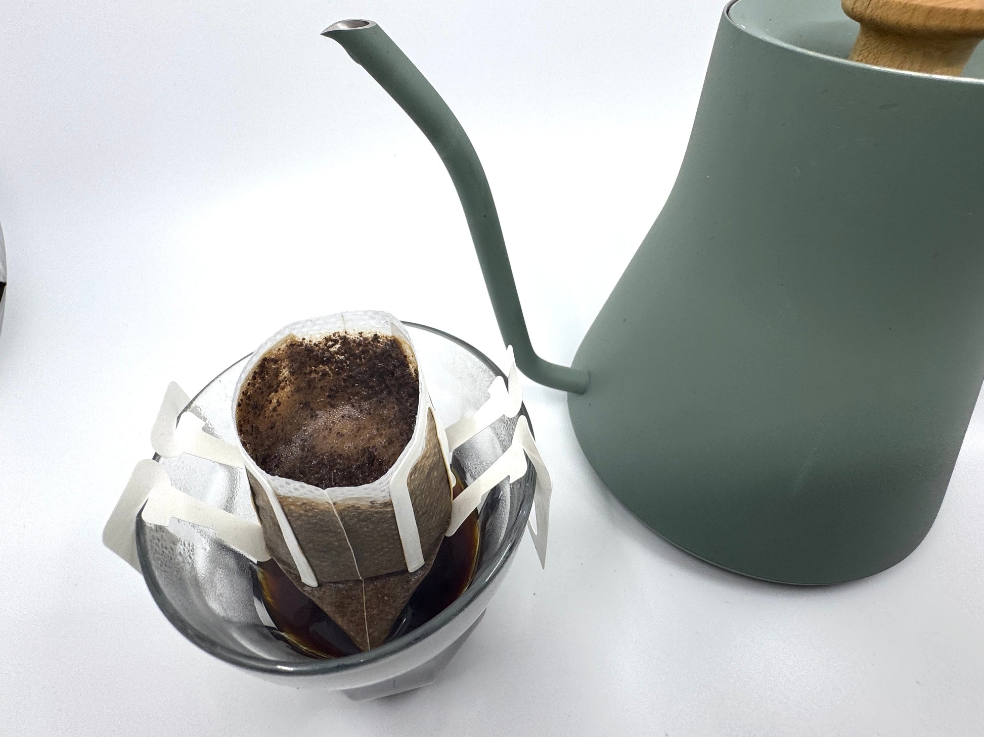 En pour-over-opsætning viser en glasdryp og kaffegrums, hvilket tyder på EverBeans Single Serve, nr. 03 · Burundi: kaffe med blomster- og bærnoter. En grøn kedel med et træhåndtag venter i nærheden for at skabe den perfekte bryg mod en almindelig hvid baggrund.