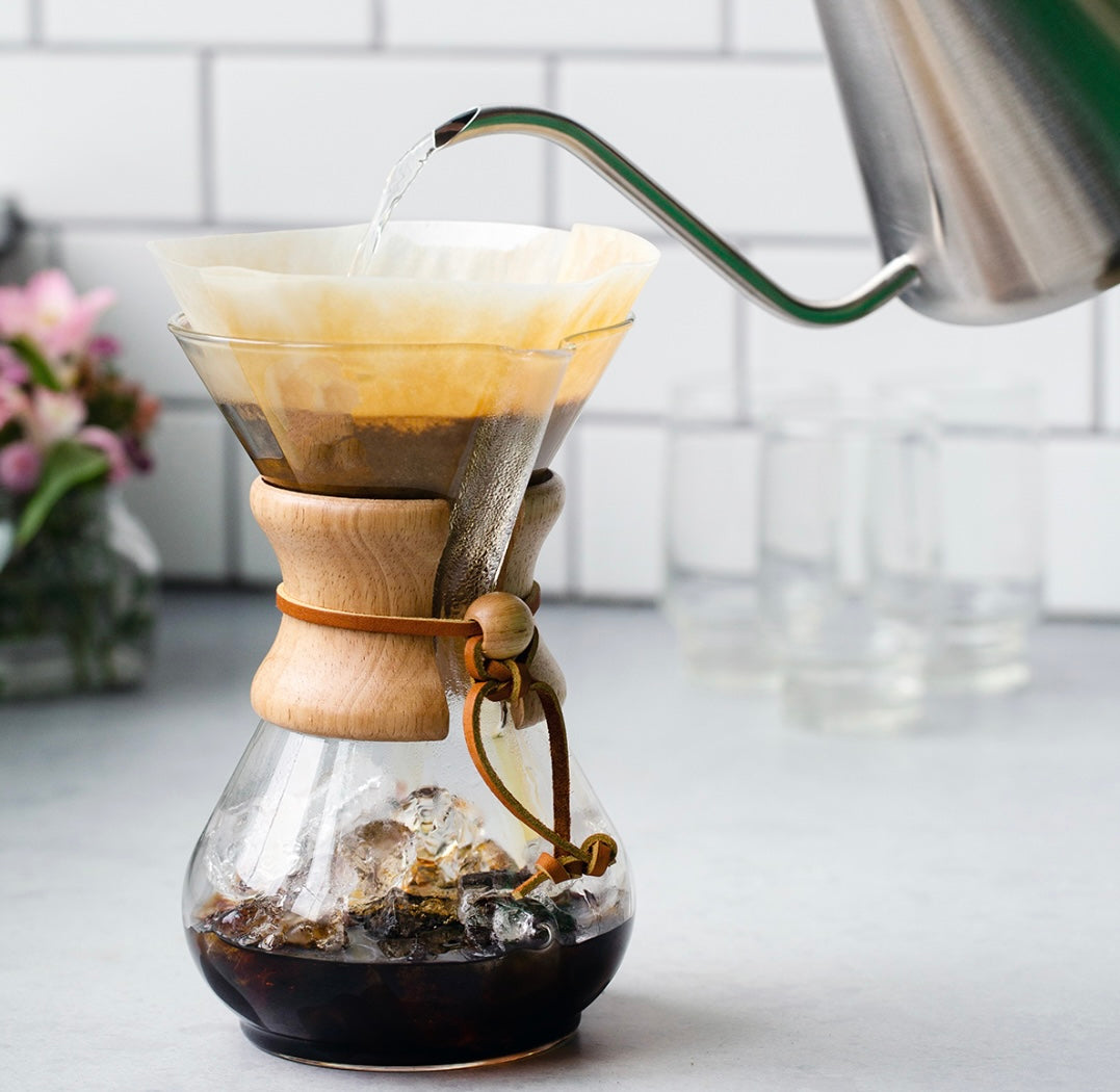 Chemex Classic Kaffebrygger 6-kopper (CM-6A) med en trækrave vugger den rige kaffe, mens vandet flyder fra en svanehals kedel, sat på baggrund af hvide fliser, delikate glas og en vase med lyserøde blomster til en fredfyldt brygningsscene .