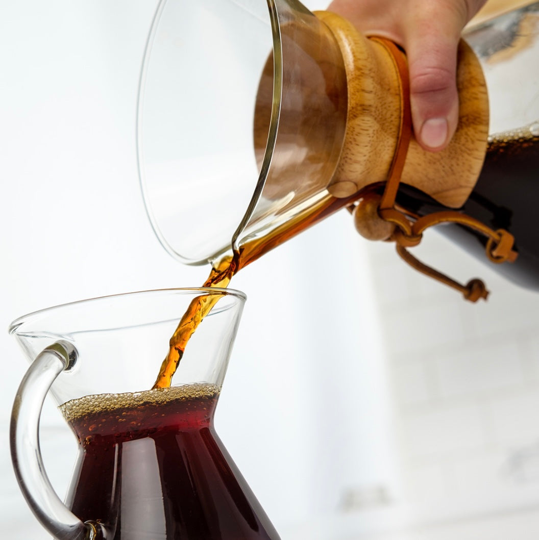 En person hælder filterkaffe fra en Chemex, Classic Kaffebrygger 6-kopper (CM-6A), i et glaskrus. Både Chemex og krus er gennemsigtige, hvilket fremhæver den mørke kaffe mod en lys, sløret baggrund.