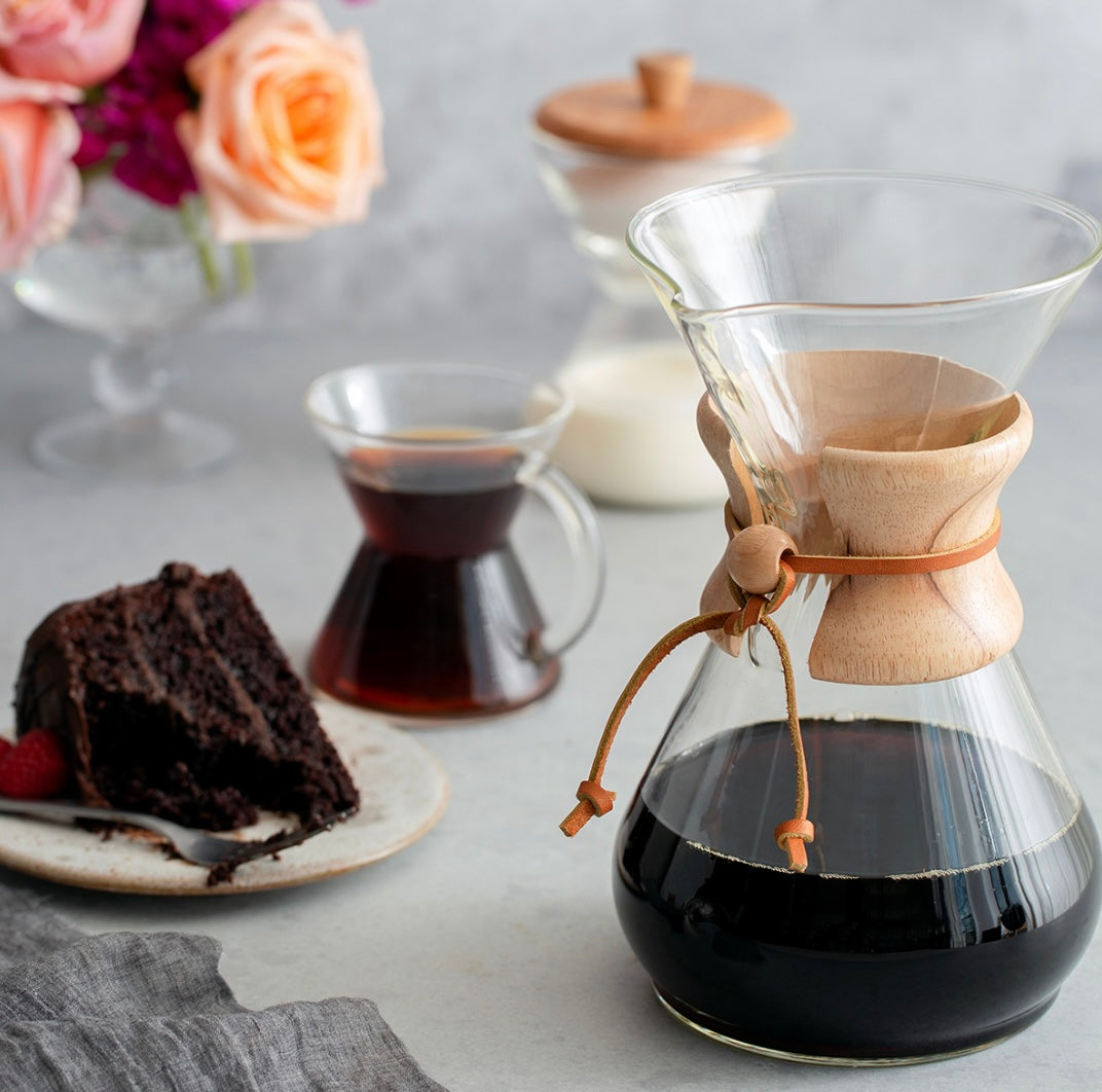 En Chemex Classic Kaffebrygger 8-kopper fra Chemex, ideel til pour-over kaffe, er elegant fyldt med rig filterkaffe på et bord. I nærheden står chokoladekage med hindbær ved siden af mælk og en vase med lyserøde roser og lilla blomster. En lille kaffekop fuldender scenen.