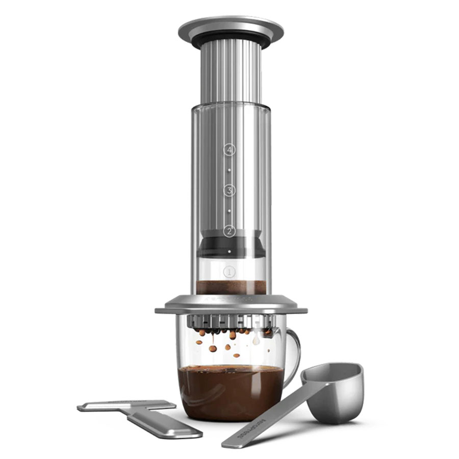 Aeropress, Premium fra Aeropress er vist med et klart kammer og tryk over et glaskrus, der samler brygget kaffe, ledsaget af en metalske og et røreværktøj, alt sammen sat mod en hvid baggrund.