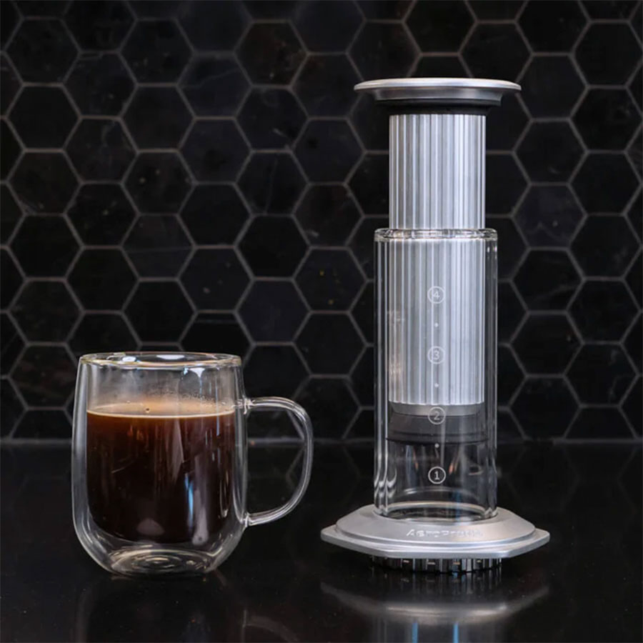 Et glaskrus kaffe står ved siden af en Aeropress Premium kaffemaskine fra Aeropress på en mørk overflade, sat mod en sort sekskantet flisevæg i baggrunden.