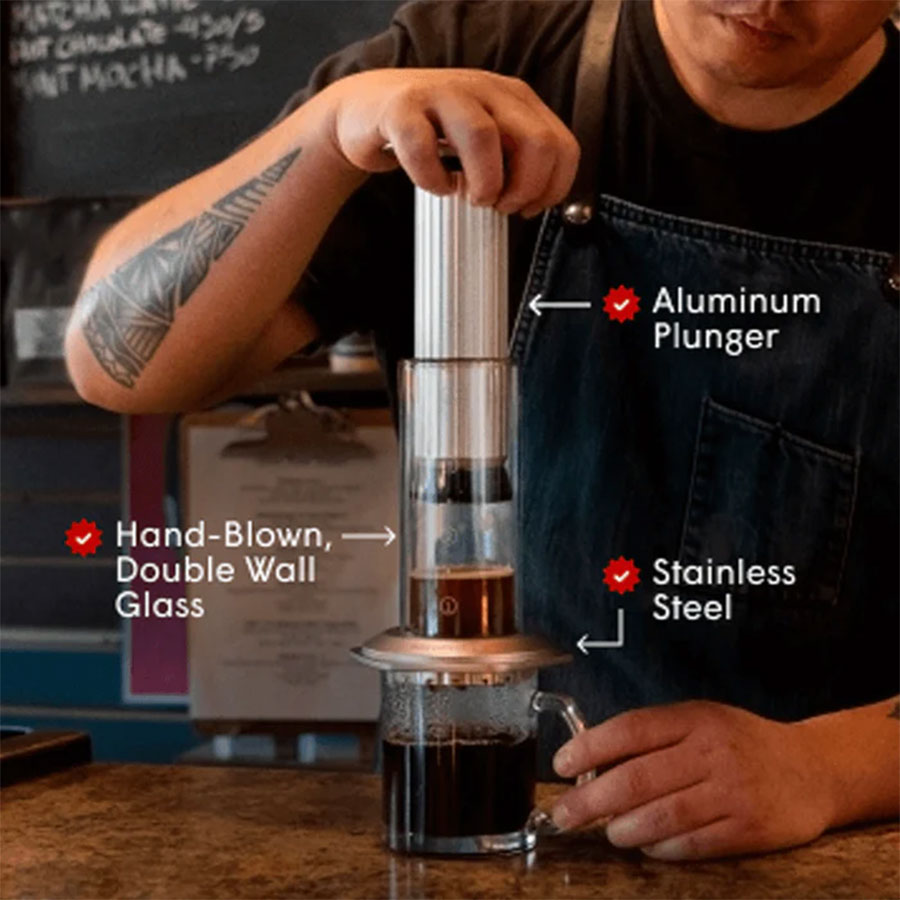 En person bruger en Aeropress, Premium med dele mærket "Aluminium Plunger", "Hand-Blown, Double Wall Glass" og "Stainless Steel", mens han er iført en tatoveret arm og mørkt forklæde.