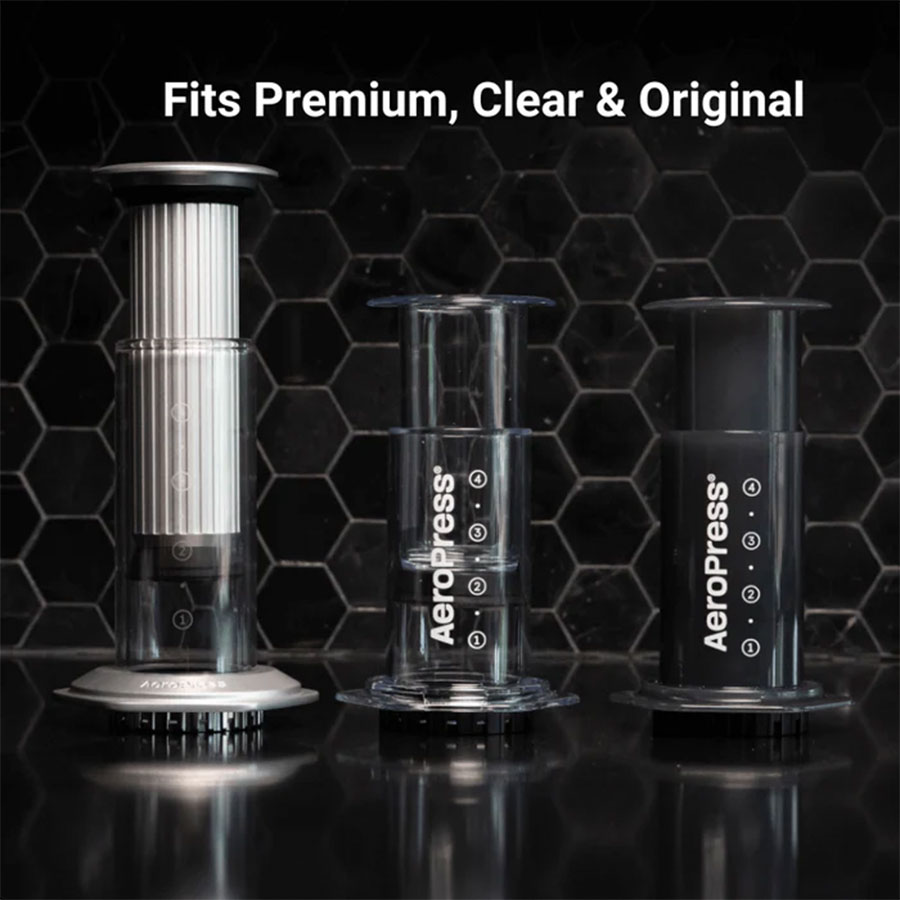 Tre Aeropress kaffemaskiner på en mørk sekskantet fliseoverflade, hver med et lidt forskelligt design: Premium, Clear og Original. Teksten ovenfor lyder "Passer til Premium, Clear & Original.