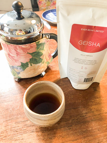 En fransk presse med en blomsterhygge sidder på et bord ved siden af en pose mærket "EverBean's Limited, Columbia Geisha", og antyder dens frugtige aroma. En lille keramisk kop rummer sort kaffe, mens en blå og hvid underkop tilføjer charme i baggrunden til dette specielle kaffeøjeblik.