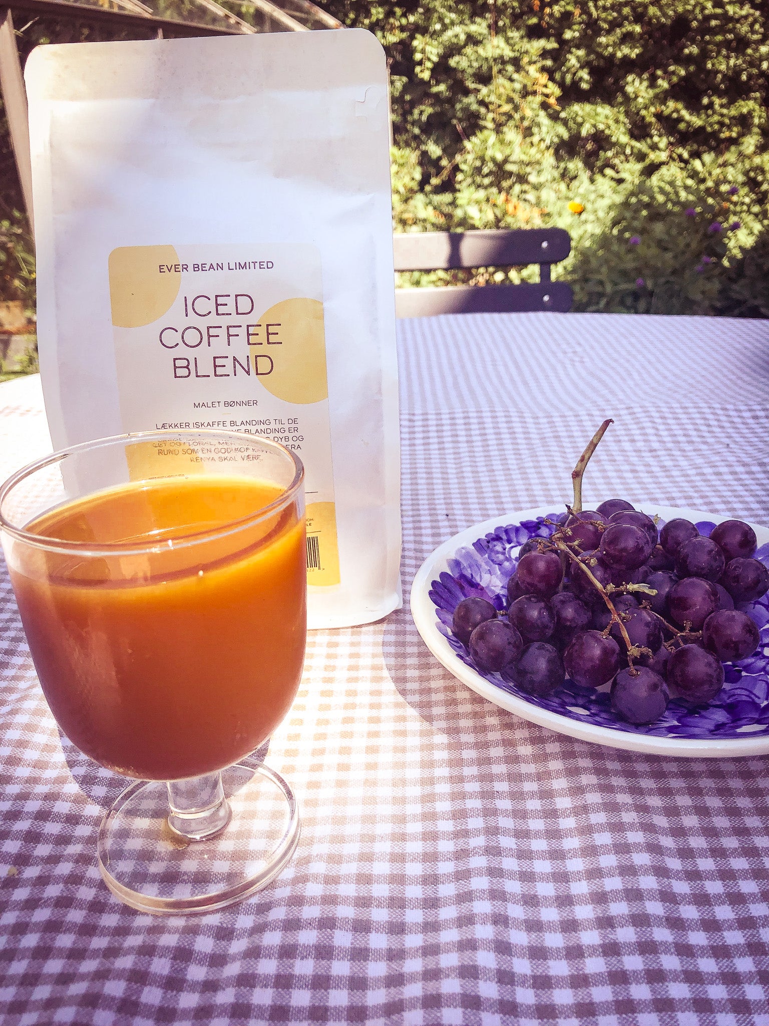 Et glas iskaffe hviler på en ternet dug ved siden af lilla druer. I baggrunden antyder en pose mærket "ICED Coffe med brygger" fra EverBean det rige bryg indeni. De udendørs omgivelser byder på smukt sløret grønt i det fjerne.