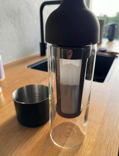 En EverBean ICED Coffe med brygger, en gennemsigtig glas te-infuser med sort låg, ideel til gennemvædning af iskaffeblandinger, hviler på en træbordplade ved håndvasken. Dens kurv er fyldt med pulveragtigt indhold. En sort metalkop er i nærheden, da slørede køkkenelementer tjener som baggrund.