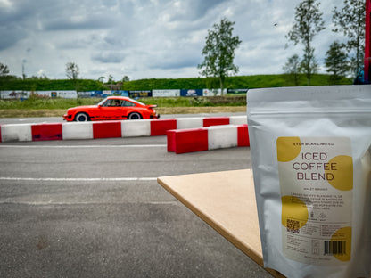 En ICED Coffe med brygger taske fra EverBean hviler på et bord nær en racerbane. En rød bil zoomer forbi, med træer og overskyet himmel i baggrunden. Røde og hvide barrierer langs banen, skaber en spændende scene – ideel til at nyde iskaffe på en spændende dag.