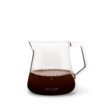 The Fellow Mighty Small Glas Karaffel, halvt fyldt med kaffe og bobler, passer perfekt sammen med Stagg [X] Drypperens for et ideelt kaffe-til-vand forhold. Den har en bred tud, robust håndtag og "Fellow" i hvid på en sort base.