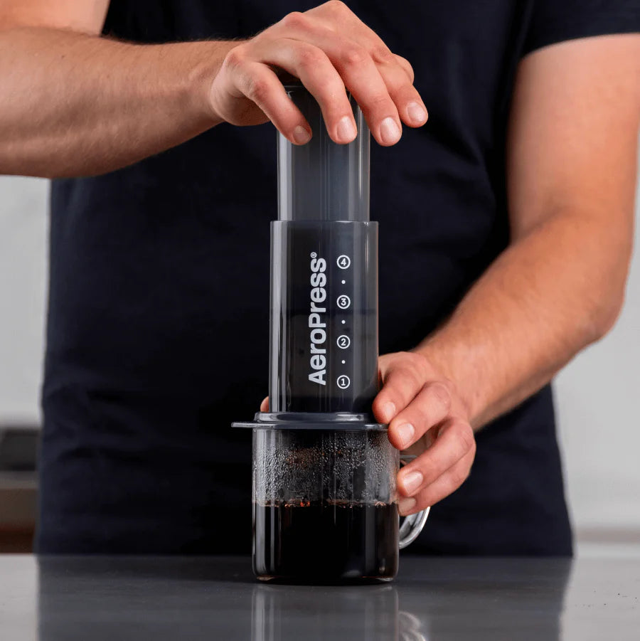 En person laver en kaffeoplevelse med en AeroPress, Original kaffebrygger fra Aeropress, der presser stemplet over et klart glas krus fyldig kaffe iført en mørk kortærmet skjorte. Den smukt slørede neutrale baggrund fremhæver bryggekunsten.