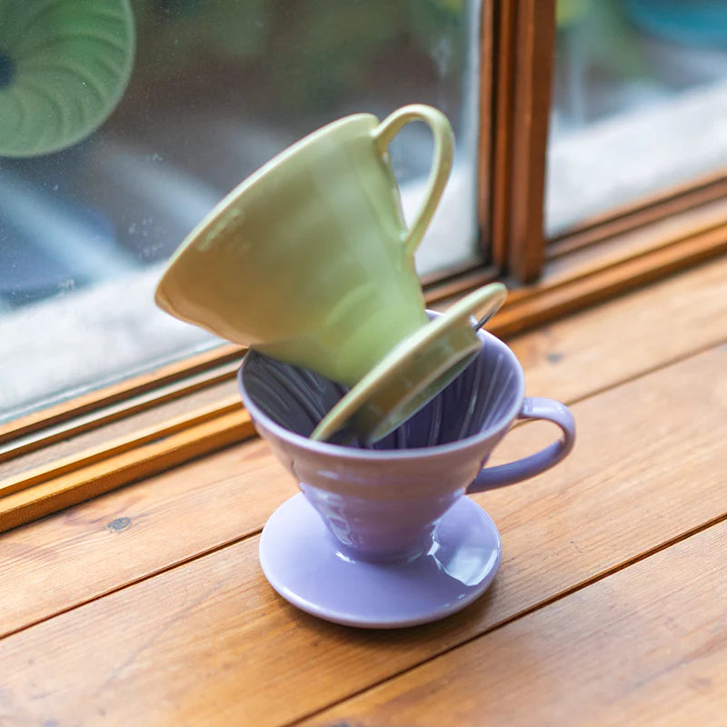 To Hario Keramisk Drip Tragt kaffedrypper, den ene i pastelgrøn og den anden i lavendel, er stablet ved et vindue på en træoverflade. Lyset skinner igennem og fremhæver deres bløde farver designet til perfekt kaffeekstraktion.